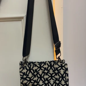 Svartvit axelväska med mönster - Snygg axelväska med justerbar svart rem och ett unikt svart mönster på vit bakgrund. Väskan har en rektangulär form och är perfekt för att bära dina viktigaste grejer. Materialet känns mjukt och har en textil känsla. Passar dig som gillar stilrena detaljer.