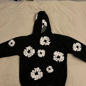 Denim tears hoodie - Svart hoodie med stora och små vita blommiga tryck över hela tröjan, inklusive huvan. Tröjan har huva och klassisk känguruficka framtill. Perfekt för dig som gillar streetwear med unik design och vill sticka ut lite extra.