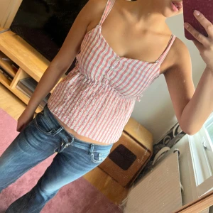 Randig rosa och vit linnetopp - Säljer en söt linnetopp med smala axelband och knappar framtill. Toppen har rosa och vita ränder, elastisk midja och är croppad i modellen. Perfekt för varma dagar och har en lätt, luftig känsla. Materialet känns som bomull och toppen är ärmlös.