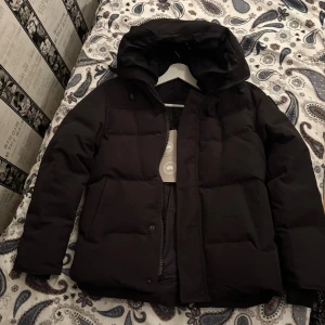Svart pufferjacka från Canada Goose - Svart pufferjacka från Canada Goose med huva och diskret logga på ärmen. Jackan har dragkedja och knappar framtill, två stora fickor och quiltad design. Fodrad med dun för extra värme. Säljs PGA för liten för mig(den är S och jag behöver M)