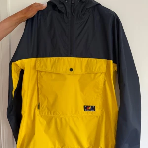 Gul och svart vindjacka från Timberland - Snygg gul och svart vindjacka från Timberland med stor ficka framtill, huva och dragkedja upptill. Jackan är vattentät och har cool logga på bröstet. Perfekt för dig som vill sticka ut med färg och stil när det blåser eller regnar.