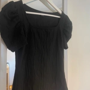 Svart blus med puffärm  - Säljer en svart blus med puffiga korta ärmar och fyrkantig halsringning. Toppen har en lätt skrynklig struktur i tyget och sitter snyggt med normal passform. Perfekt för dig som gillar stilrena och trendiga plagg.