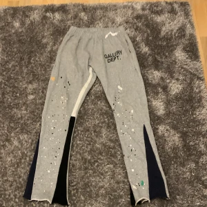 Grå mjukisbyxor från Gallery Dept. - Grå mjukisbyxor från Gallery Dept. med svart logga på ena benet. Byxorna har färgstänk i vitt, svart, grönt och gult samt svarta och vita paneler längs benen. Tillverkade i 100% bomull och har elastisk midja för skön passform.