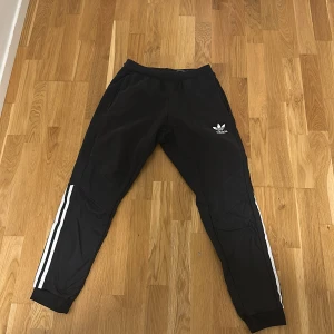 Svarta Adidas mjukisbyxor med vita ränder - Svarta mjukisbyxor från Adidas med klassiska vita ränder längs benen och Adidas logga på låret. Byxorna har ribbade muddar vid bensluten och är gjorda i ett mjukt syntetmaterial. Perfekta för chill eller träning.