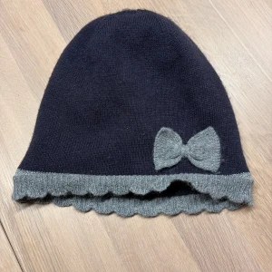 Mörkblå mössa med rosett Polarn O. Pyret - Supersöt mörkblå mössa från Polarn O. Pyret med ljusgrå kant och en matchande rosett framtill. Mössan har vågig ribbad kant och är tillverkad i en mjuk blandning av nylon, polyester och viskos. Perfekt för kyliga dagar.