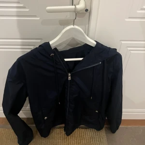 Mörkblå vindjacka från Moncler - Snygg mörkblå vindjacka från Moncler med huva och dragsko. Jackan har dragkedja framtill, två fickor med tryckknappar och Moncler-logga på ärmen. Röd och vit randig detalj på huvan ger en sportig vibe. Perfekt för vår och höst.