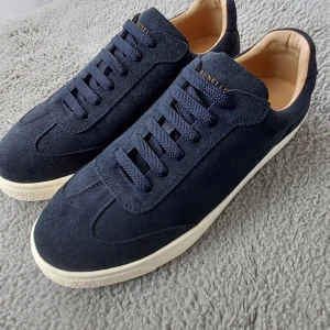 Mörkblå sneakers från Brunello Cucinelli - Snygga mörkblå sneakers i mocka från Brunello Cucinelli med vita gummisulor och klassisk snörning. Skorna har en stilren design med rund tå och beige innersida i skinn. Perfekta för dig som gillar exklusiva och tidlösa sneakers.