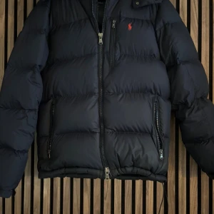 Mörkblå pufferjacka från Polo Ralph Lauren - Säljer en mörkblå pufferjacka från Polo Ralph Lauren med röd logga på bröstet. Jackan har huva, två sidofickor med dragkedja och justerbar nederkant med snörning. Perfekt för kalla dagar och har en stilren look.