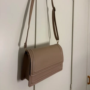 Beige axelväska i skinnimitation - Stilren beige axelväska i skinnimitation med justerbart axelband och magnetlås i guld. Väskan har en fyrkantig form och minimalistisk design, perfekt för att bära det viktigaste. Snygga detaljer med markerade sömmar och enkel insida.  Shien 