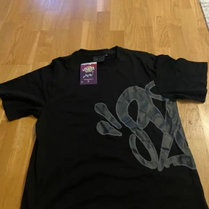 Svart och grå syna world set. - Svart t-shirt och shorts från Syna world med stort grått graffiti-inspirerat tryck både fram och bak. Klassisk passform med rund hals och korta ärmar. Perfekt för dig som gillar streetwear och vill sticka ut med en unik design.