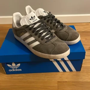 Adidas Gazelle grå/vita sneakers - Säljer ett par klassiska Adidas Gazelle sneakers i grå mocka med vita detaljer och vita ränder på sidorna. Skorna har snörning och en platt sula, samt Adidas-logga på plösen och hälen. Perfekta för dig som gillar retrostil och streetwear.De fins också ett par vita snören till.