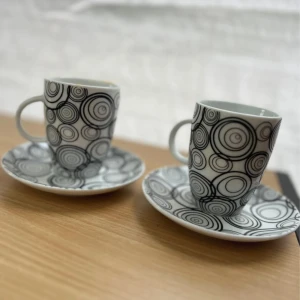 Set med 2 kaffekoppar med fat, svartvitt cirkelmönster - Två stilrena kaffekoppar med tillhörande fat i porslin. Unikt svartvitt cirkelmönster ger en modern känsla. Koppar och fat är i fint skick utan synliga sprickor eller nagg. Perfekt för kaffe eller espresso och passar både till vardag och fest.