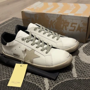 Golden Goose vita sneakers med stjärna - Golden Goose sneakers i vitt skinn med grå mockastjärna på sidan, grå snörning och svart hälparti. Klassisk låg modell med platt sula och diskreta detaljer. Snygg och stilren design som passar till det mesta. Levereras med originalkartong och tag.