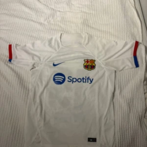 FC Barcelona vit bortatröja Lamine Yamal - Säljer en vit FC Barcelona bortatröja från Nike med Lamine Yamal och nummer 19 på ryggen. Tröjan har korta ärmar med röda och blå detaljer, klubbmärke på bröstet och Spotify-logga framtill. Materialet är lätt och andas, perfekt för fotboll eller träning. Storlek 170 cm skick ny utan prislapp 