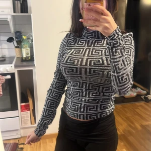 Glittrig polotopp från New Yorker  - Säljer en glittrig polotopp från New Yorker i svart och silver med ett grafiskt mönster. Toppen är långärmad, tight och har en hög krage. Materialet är genomskinligt med glittertrådar som ger en riktigt snygg effekt. Perfekt för dig som vill sticka ut!