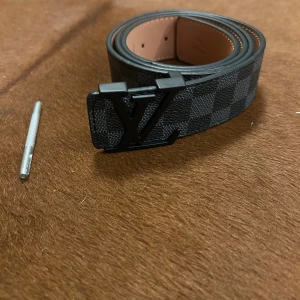 Louis Vuitton Damier svart bälte - Snyggt Louis Vuitton-bälte med klassiskt svart och grått rutmönster, ikoniskt LV-spänne i matt svart metall och insida i ljust skinn. Perfekt accessoar för att lyfta din outfit med exklusiv känsla och stil.