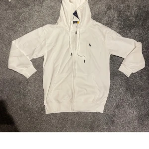 Vit hoodie från Polo Ralph Lauren M - Vit hoodie från Polo Ralph Lauren med dragkedja, snörning i huvan och klassisk logga på bröstet. Tröjan har långa ärmar, ribbade muddar och två fickor framtill. Perfekt för en chill och stilren look.