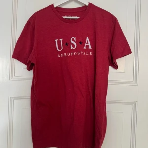 Röd t-shirt med tryck - Röd t-shirt i storlek L med vit och svart USA-tryck på bröstet. Klassisk rund halsringning och korta ärmar. Skön och mjuk bomullskänsla, perfekt för en avslappnad stil.