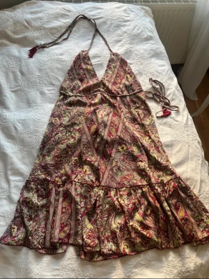 Paisleymönstrad långklänning med knyt - Paisleymönstrad långklänning i vinröda, gula och beige toner med blommiga detaljer. Klänningen har omlott-v-ringad hals, knyt i nacken och volang nedtill. Materialet är 80% konstsilke och 20% polyester, vilket ger ett mjukt och glansigt fall. Köptes på en marknad i Italien förra året 