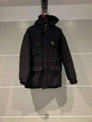 Svart dunjacka från Fjällräven - Svart dunjacka från Fjällräven med huva och flera stora fickor framtill. Jackan har dragkedja och klassisk Fjällräven-logga på bröstet. Yttertyg i nylon, fodrad med nylon och polyester, fylld med 80% dun och 20% fjäder. Perfekt för kalla dagar.