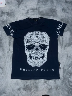 Navyblue Philipp Plein t-shirt med döskalle - M men passar S. Mörk blå Philipp Plein med stor vit döskallegrafik glitter på och tryck på ärmarna. Klassisk passform med rund hals och coola detaljer. Materialet känns mjukt och skönt, perfekt för dig som gillar streetstyle och statement-plagg.