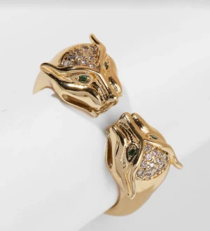 Guldfärgad panterring - Cool ring i guldfärg med två panterhuvuden som möts framtill. Huvudena har gröna ögon och är dekorerade med glittriga stenar på ovansidan. Ringen har en unik och edgy design som verkligen sticker ut och passar dig som gillar djurmotiv och detaljer.