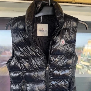 Svart dunväst från Moncler - Snygg svart dunväst från Moncler med glansig finish och klassisk logga på bröstet. Västen har hög krage, dragkedja framtill och quiltad design. Perfekt för lager-på-lager och ger en cool vibe till din outfit. BILLIGARE VID SNABB AFFÄRR!!