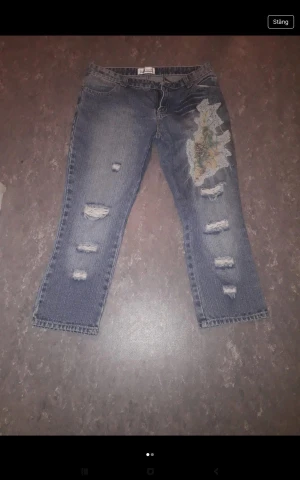 Blå jeans med broderad patch - Säljer ett par blå jeans med slitna detaljer och en stor broderad patch med blommönster på ena benet. Jeansen har rak passform och flera hål för en cool, edgy look. Perfekt för dig som gillar unik stil och detaljer.
