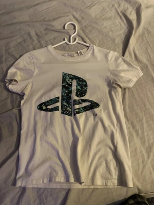 PlayStation t-shirt från H&M - Vit t-shirt från H&M med stort PlayStation-tryck i blått och svart med coolt splattermönster på bröstet. Klassisk rund hals och korta ärmar, perfekt för dig som gillar gaming och vill ha en chill stil. Gjord i mjuk bomull för skön känsla hela dagen.