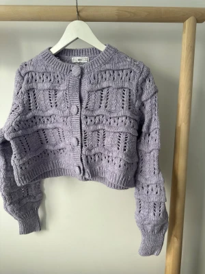 Ljuslila stickad kofta från MANGO - Superfin ljuslila kofta från MNG i stickad ullblandning. Croppad modell med långärmade ärmar och snyggt mönster över hela koftan. Stora matchande knappar framtill och ribbade muddar vid hals, ärmslut och nederkant. 