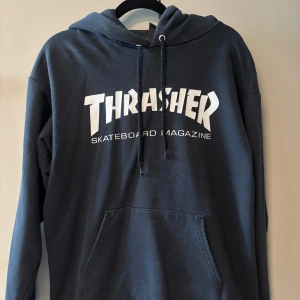Svart Thrasher hoodie med tryck - Svart hoodie från Thrasher med stort vitt tryck på bröstet och klassisk magficka. Tröjan har huva med snörning och är långärmad, perfekt för skate eller chill. Mjuk insida och cool streetstil, märkt med Thrasher San Francisco. Tvättats några gånger men är fortfarande i väldigt bra skick