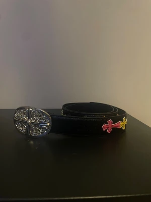 Svart skärp med kors och silverdetalj - Snyggt svart bälte i skinn med en stor, oval silverspänne med detaljerat mönster. Bältet har broderade kors i rosa och gult på sidan som ger en unik och cool vibe. Perfekt accessoar för att lyfta din outfit.