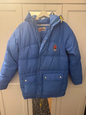 Blå Fjällräven dunjacka med huva - Säljer en blå dunjacka från Fjällräven med huva, två stora fickor med knappar och gul snörning nedtill. Jackan har broderad Fjällräven-logga på bröstet och är quiltad för extra värme. Perfekt för kalla dagar och har en loose passform.
