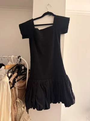 Svart kort klänning med volang - Supersöt svart kort klänning med fyrkantig ringning och puffiga korta ärmar. Överdelen är smockad och kjolen har en volangig, bubblig effekt som ger extra volym. 