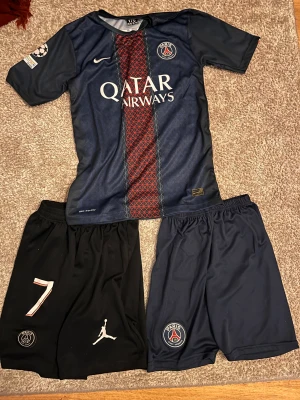 PSG shorts svart & marinblå, stl 176 S passar bra för stl 164 - Två par PSG shorts säljes! Ena paret är svart med nummer 7, Jordan-logga och PSG-märke, andra paret är marinblå med PSG-märke. Båda har resår i midjan och är perfekta för fotboll eller träning. Snygg sportig look och officiella klubbmärken.