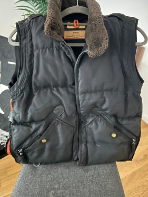 Parajumpers väst - Svart dunväst från Parajumpers med bruna detaljer i kragen och flera praktiska fickor med dragkedja och knappar. Västen har broderade märken på ryggen och insidan, samt en mjuk, fluffig krage. Perfekt för kalla dagar och har en cool, urban look.