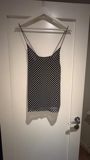 Svart prickig topp med tunna band - Supersöt svart topp med vita prickar och tunna axelband. Toppen har en rak passform och ärmlös design, perfekt för varma dagar. Materialet känns lätt och svalt mot huden. Prickmönstret ger en lekfull och trendig vibe.