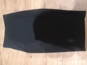 Svart pennkjol från AMISU, storlek 34 - Svart, stilren pennkjol från AMISU i storlek 34. Kjolen har en klassisk rak form och är knälång, perfekt för en clean och snygg look. Materialet känns mjukt och har en lätt stretch, vilket gör den bekväm att bära. Passar dig som gillar minimalistisk stil. Kjolen är lite noppig på baksidan. Se sista bild. 