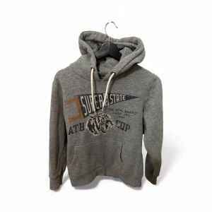 Grå Superdry hoodie med tryck - Snygg grå hoodie från Superdry med coolt tryck på bröstet och en tiger. Tröjan har huva med vita snören, känguruficka och tryck i blått och orange på ärmen. 