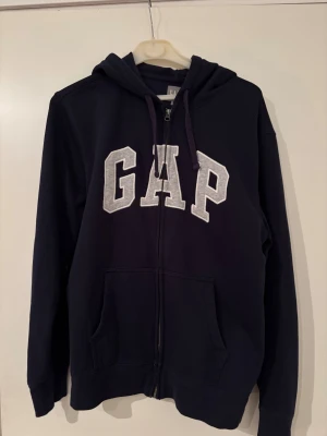  Hoodie GAP  - Säljer en mörkblå hoodie från GAP i storlek L. Tröjan har vit GAP-logga framtill, huva med snörning och praktiska fickor. Materialet är mjuk bomull och den har lång ärm samt dragkedja hela vägen 