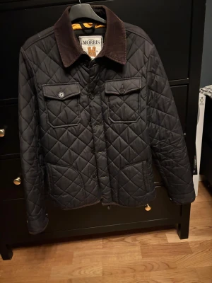 Svart quiltad jacka från Morris M - Quiltad svart jacka från Morris med bruna manchesterkrage, guldfärgad broderad logga på ärmen och två bröstfickor med knappar. Jackan har snyggt rutmönster och guldfärgat innerfoder. Perfekt för dig som gillar klassisk stil med modern touch.