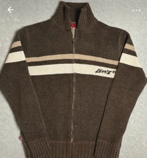 Brun stickad kofta från Levi's - Snygg brun stickad kofta från Levi's med dragkedja och högt ribbat krage. Tröjan har vita och beige ränder över bröstet och armarna, samt Levi's-logga på framsidan. Långärmad med breda muddar och mjukt material som håller dig varm.