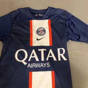 PSG Mbappé matchtröja Nike blå - Paris Saint-Germain fotbollströja med Mbappé 7 på ryggen. Djupblå med vita och röda detaljer, PSG-logga och Qatar Airways-tryck på bröstet. Tillverkad i lätt och ventilerande material, perfekt för fotboll eller att bära som supporter.