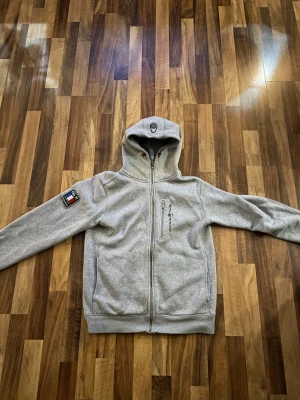 Grå hoodie från Sail Racing FRA 77 - Grå hoodie från Sail Racing med FRA 77-tryck på ryggen och logga på bröstet. Tröjan har hel dragkedja, huva med snörning och en bröstficka med dragkedja. Materialet är mjuk bomull/polyesterblandning och passformen är normal. Snygga röda detaljer vid snörningen.