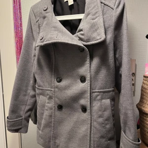 Grå kappa från H&M, stl 36 - Snygg grå kappa från H&M i klassisk modell med dubbelknäppning och bred krage. Jackan har två fickor framtill. 