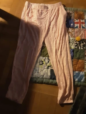 Ljusrosa mjukisbyxor med resår - Säljer ett par ljusrosa mjukisbyxor med skön resår i midjan. Byxorna har ett enkelt och avslappnat snitt, perfekta för chill dagar hemma. Materialet är mjukt och lätt, och färgen är diskret rosa med svaga rutor i mönstret.