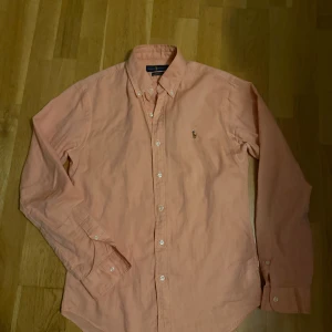 Rosa skjorta från Ralph Lauren S - Snygg rosa skjorta från Ralph Lauren i slim fit med klassisk button down-krage och broderad logga på bröstet. Skjortan är i bomull och har långa ärmar samt vita knappar framtill. 