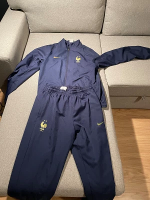 Nike Frankrike fotbollsträningsoverall - Mörkblå träningsoverall från Nike med Frankrikes landslagsemblem och guldiga detaljer. Setet består av jacka med dragkedja och byxor med resår i midjan. Tillverkad i Dri-FIT polyester som andas, perfekt för fotbollsträning.