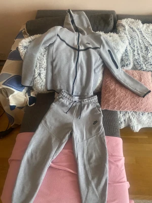 Nike tech set grå - Snygg grå hoodie från Nike med svart dragkedja och huva. Tröjan har lång ärm, en ficka med dragkedja på armen och svarta detaljer längs bröstet. Perfekt för chill eller träning, matcha med dina favoritjoggers för en clean look.