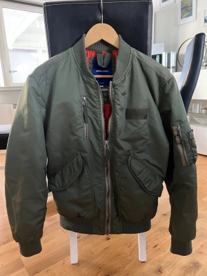 Olivgrön bomberjacka Jack & Jones S - Snygg olivgrön bomberjacka från Jack & Jones med orange foder, ribbade muddar och flera fickor med dragkedja. Jackan är vattenavvisande och har återvunnet foder. Perfekt för dig som gillar streetstyle och vill ha en klassisk look.